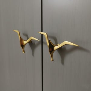 May include: Two gold-colored cabinet handles in a bird-like design are mounted on a gray cabinet door. The handles have a modern, minimalist aesthetic, with a sleek, angular form. The handles are made of a shiny, reflective material.