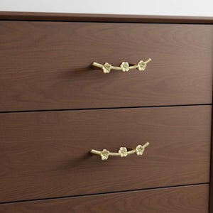 May include: A dark brown wooden chest of drawers with two visible drawers. Each drawer is fitted with a gold-coloured floral branch handle. The handles are curved and feature small flower details, adding a touch of elegance.