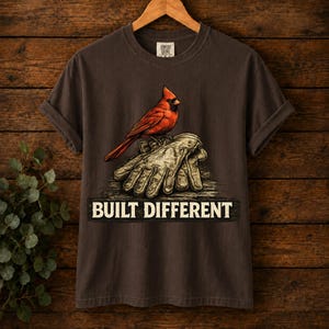 Può includere: Una t-shirt a maniche corte marrone scuro con l'illustrazione di un cardinale rosso brillante appollaiato su un paio di guanti da lavoro color cuoio. Sotto i guanti, la scritta "BUILT DIFFERENT" è stampata in un carattere bianco sporco.