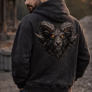 May include: Black hoodie featuring a detailed illustration of a ram's head with glowing orange eyes. The artwork is centered on the back of the hoodie. The hoodie is a dark gray color and has a drawstring hood.