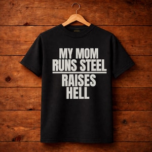 Puede incluir: Camiseta negra con el texto blanco "MY MOM RUNS STEEL RAISES HELL". La camiseta está colgada en una percha de madera sobre un fondo de madera. El texto está en negrita, fuente sans-serif.