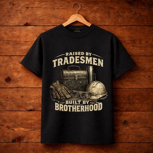 May include: A black short-sleeve t-shirt featuring a graphic print of a metal toolbox, a thermos, work gloves, safety glasses, and a hard hat. The text reads RAISED BY TRADESMEN and BUILT BY BROTHERHOOD in a beige, distressed, block-style font.