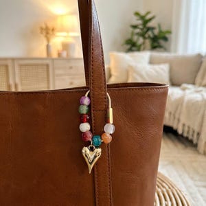 Op de afbeelding: Een bruine leren tote bag met een goudkleurige hartbedel en een kralenaccent. De bedel heeft een gouden hartvormige hanger en kleurrijke kralen in verschillende kleuren, waaronder paars, groen, rood en oranje.