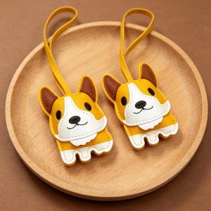 May include: Two yellow, white, and brown corgi-shaped keychains with yellow straps. The keychains are made of stitched leather and have a smiling face design. The keychains are displayed on a wooden plate.