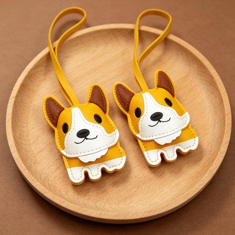 May include: Two yellow, white, and brown corgi-shaped keychains with yellow straps. The keychains are made of stitched leather and have a smiling face design. The keychains are displayed on a wooden plate.