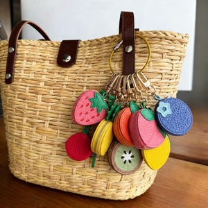 May include: A woven straw tote bag with brown leather handles and a decorative fruit-themed keychain. The keychain features miniature zippered pouches shaped like a strawberry, cherry, kiwi, lemon, orange, and blueberry.