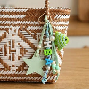 May include: A woven brown and white basket bag featuring a decorative charm attached to the handle. The charm includes a light green star, a small crocheted green fish, and letter beads spelling HBA in white, green, and blue, accented with pearls.