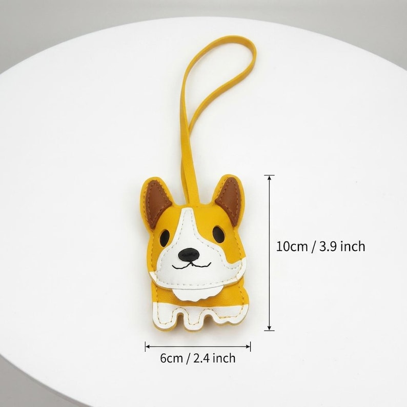 May include: A yellow, white, and brown corgi-shaped keychain with a loop. The keychain is 3.9 inches tall and 2.4 inches wide. The corgi has brown ears, a white face, and a smiling expression.