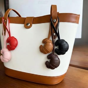 May include: A white and tan tote bag with a brown handle and clasp. Several croissant-shaped keyrings hang from the handle in shades of pink, red, light brown, dark brown, and black. The bag has a white body and tan trim.