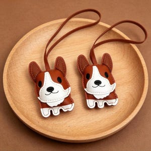May include: Two brown and white corgi-shaped keychains with brown straps. The keychains are made of leather and have black eyes and noses. They are resting on a wooden plate.