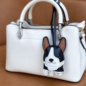 May include: A white handbag with a top handle and silver hardware. A black, white, and brown dog-shaped charm hangs from the handle. The charm has a smiling face and is made of leather.