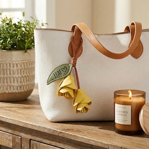 Può includere: Una borsa tote in pelle bianca con manici in pelle color cuoio e un ciondolo decorativo composto da una foglia verde con la scritta LEAF e un cuore, accompagnata da due nappe in pelle gialla a forma di fiore su un sottile cordino.