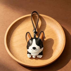 May include: A black, white, and tan corgi-shaped keychain with a black leather loop. The keychain is sitting on a wooden plate. The corgi has tan ears, a white face, and a black body.