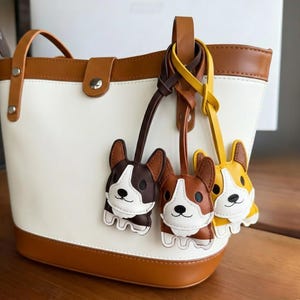 May include: A white and tan tote bag with three corgi-shaped keyrings in brown, tan, and yellow. The bag has a tan handle and trim. The keyrings are attached to the bag's handle.