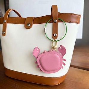 May include: White tote bag with brown trim and handles. A pink crab-shaped keyring hangs from a green wire. The bag has a structured design with a button closure.