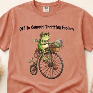 May include: A terracotta-coloured t-shirt featuring a frog on a penny-farthing bicycle, with a basket of flowers. The text "Off To Commit Thrifting Foolery" is printed above the frog. The shirt is made of a soft material.