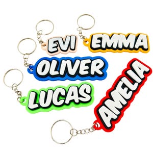 May include: A selection of name keyrings in various colours. The keyrings display the names EVI, EMMA, OLIVER, LUCAS, and AMELIA in white lettering, each with a coloured border. They are attached to silver keyrings.