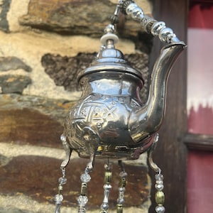 Peut inclure: Un carillon éolien en forme de théière argentée ornée, avec des brins de perles pendantes. La théière présente des motifs décoratifs et est suspendue par une chaîne. Les perles sont transparentes, argentées et dorées. La théière a de petits pieds.