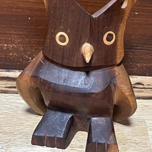 Peut inclure: Une figurine de hibou en bois fabriquée à la main, avec une finition marron bicolore. Le hibou a de grands yeux ronds avec des accents de couleur claire, un petit bec et une posture stylisée et droite. Les ailes du hibou sont arrondies et ses pieds sont carrés.
