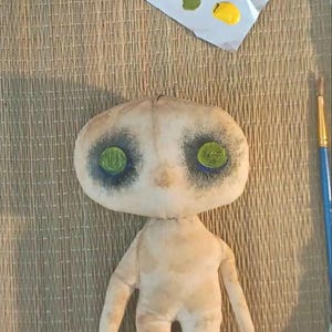 May include: A handmade doll with a pale, textured fabric body and limbs. The doll's face has large, dark eye sockets with green eyes. A palette with blue, green, yellow, and black paint sits nearby, along with a paintbrush.