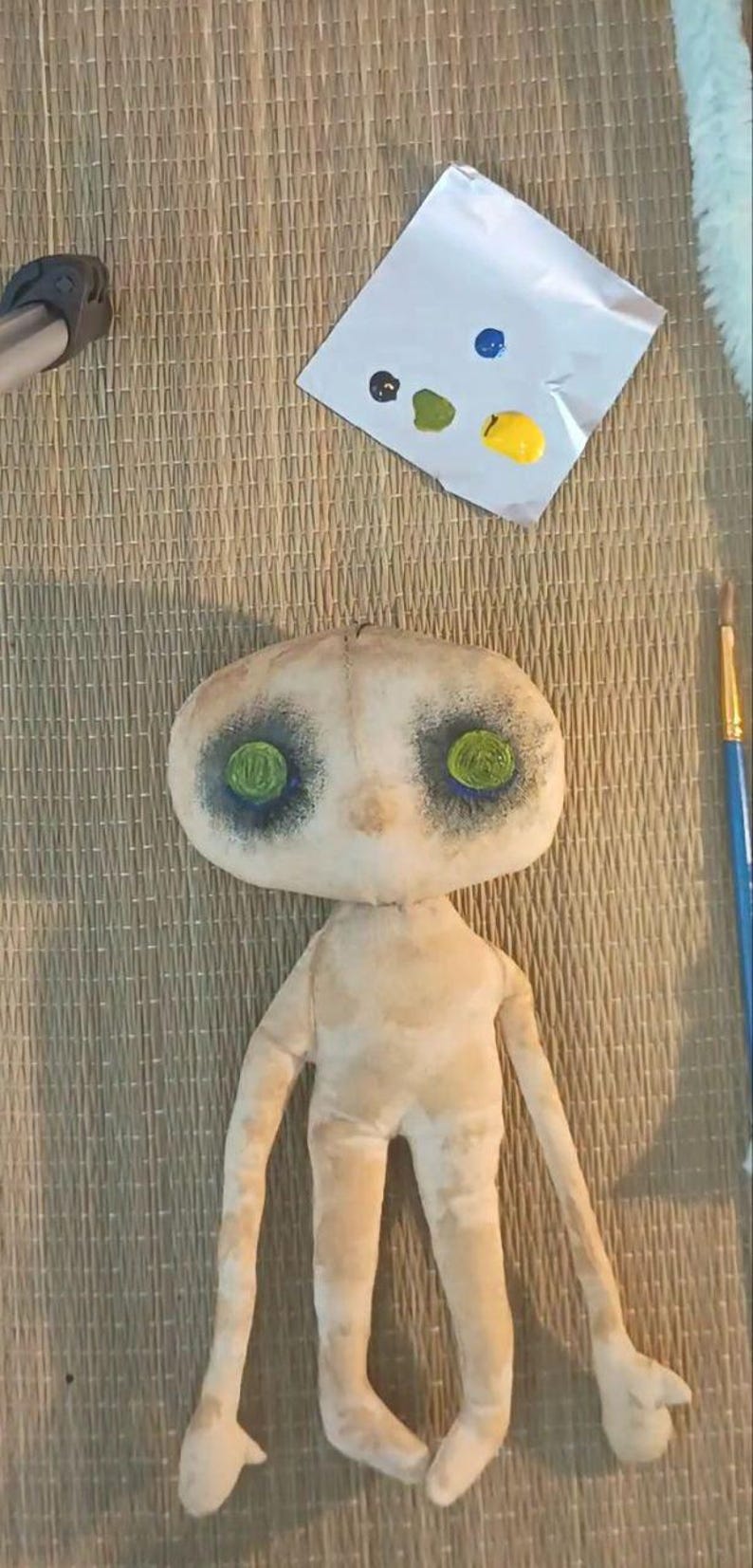 May include: A handmade doll with a pale, textured fabric body and limbs. The doll's face has large, dark eye sockets with green eyes. A palette with blue, green, yellow, and black paint sits nearby, along with a paintbrush.
