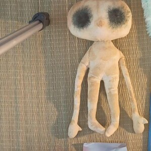 May include: A handmade doll with a large, oval head and long limbs. The doll has dark, circular eye markings and is made of a light beige fabric. The doll is lying on a woven mat.