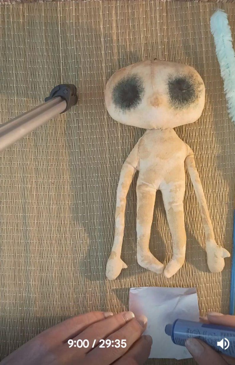 May include: A handmade doll with a large, oval head and long limbs. The doll has dark, circular eye markings and is made of a light beige fabric. The doll is lying on a woven mat.