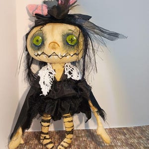 May include: A handmade doll with a pale face, green eyes, and a stitched mouth. It wears a black dress with a white lace collar, a black headpiece with a pink bow, and striped legs. The doll has a whimsical, spooky aesthetic.
