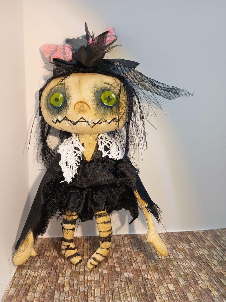 May include: A handmade doll with a pale face, green eyes, and a stitched mouth. It wears a black dress with a white lace collar, a black headpiece with a pink bow, and striped legs. The doll has a whimsical, spooky aesthetic.