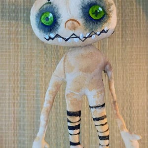 May include: Handmade fabric doll with a pale complexion, stitched details, and painted features. The doll has large green eyes, a stitched mouth, and black and white striped legs. The arms and body are a light beige color.