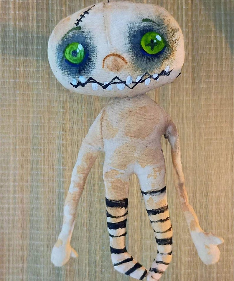 May include: Handmade fabric doll with a pale complexion, stitched details, and painted features. The doll has large green eyes, a stitched mouth, and black and white striped legs. The arms and body are a light beige color.