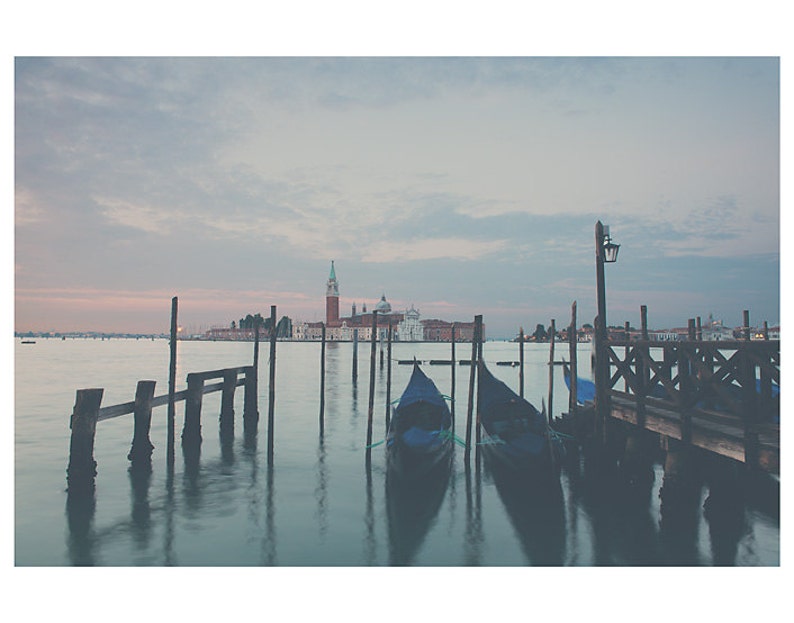 May include: A tranquil photograph of Venice, Italy, showing gondolas moored near a wooden pier. The image captures the calm water, the city's architecture, and a soft, muted colour palette, creating a sense of peace and serenity.