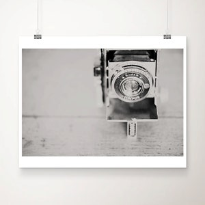 May include: Black and white photograph of a vintage Kodak camera. The camera lens is in focus, with the word "Kodak" visible. The camera is set against a light-colored wooden surface.