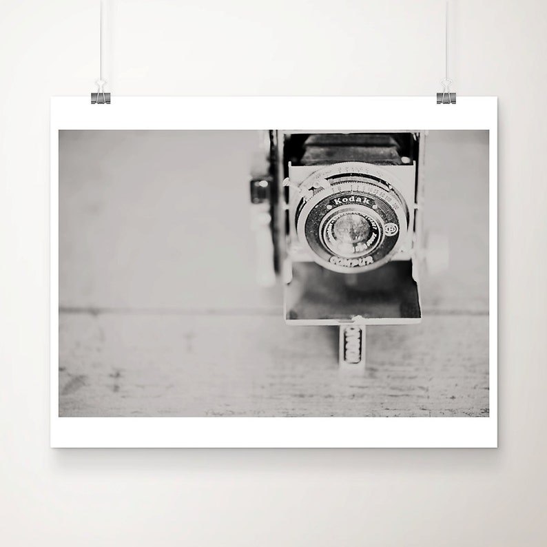 May include: Black and white photograph of a vintage Kodak camera. The camera lens is in focus, with the word "Kodak" visible. The camera is set against a light-colored wooden surface.