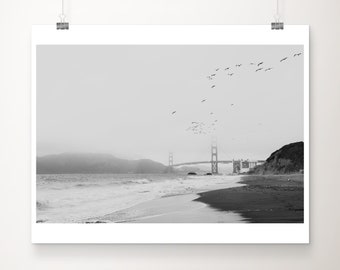 May include: Black and white print of the Golden Gate Bridge in San Francisco. The image shows the bridge in the distance, with a beach and ocean in the foreground. Birds fly in the sky above the bridge.