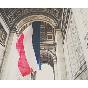 May include: A low-angle shot of the Arc de Triomphe in Paris, France, with a large French flag hanging in the center. The flag's red, white, and blue stripes are prominently displayed against the backdrop of the monument's ornate architecture.