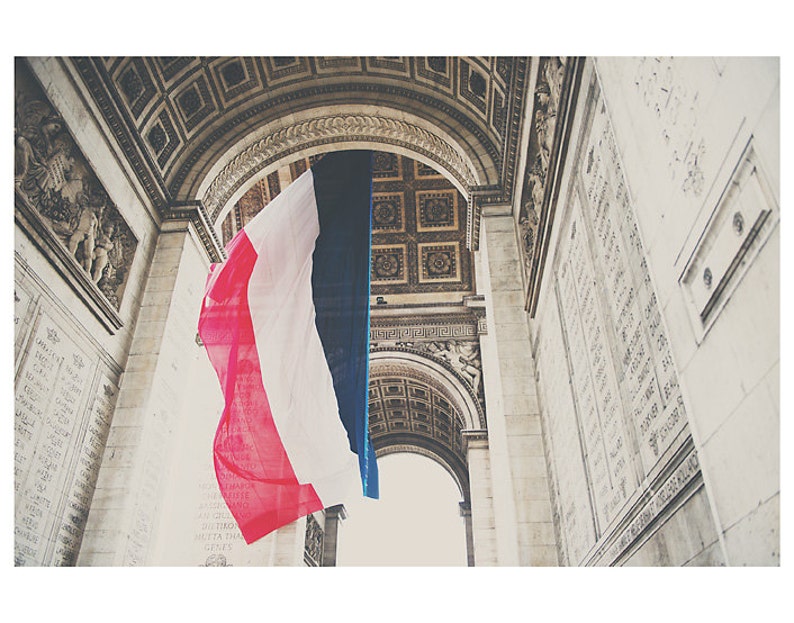 May include: A low-angle shot of the Arc de Triomphe in Paris, France, with a large French flag hanging in the center. The flag's red, white, and blue stripes are prominently displayed against the backdrop of the monument's ornate architecture.