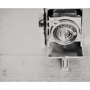 May include: Black and white vintage Kodak camera with a Compur lens. The camera is open, revealing the lens and internal components. The camera is set against a light-colored wooden surface.