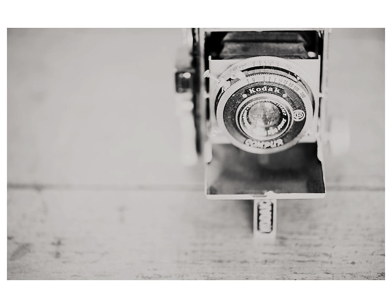 May include: Black and white vintage Kodak camera with a Compur lens. The camera is open, revealing the lens and internal components. The camera is set against a light-colored wooden surface.