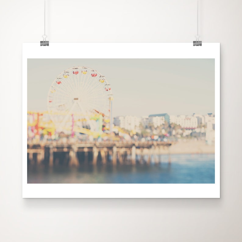 May include: A print of a blurred image of a Ferris wheel with colorful cars, a pier, and buildings in the background. The sky is a pale blue, and the water is a darker blue. The image is mounted on a white background.