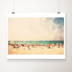 May include: A vintage-style photograph of a crowded beach with people sunbathing and swimming in the ocean. The sky is a soft blue with white clouds and a hazy, out-of-focus effect.