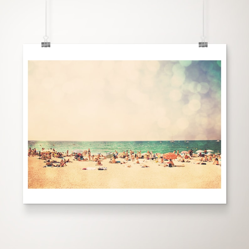 May include: A vintage-style photograph of a crowded beach with people sunbathing and swimming in the ocean. The sky is a soft blue with white clouds and a hazy, out-of-focus effect.