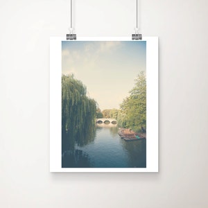May include: Framed print of a tranquil river scene. A white arched bridge crosses the water, framed by green trees and a weeping willow. The water reflects the sky, creating a peaceful and picturesque landscape.