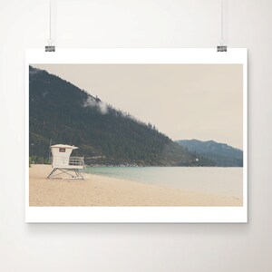 May include: A scenic landscape print featuring a white lifeguard stand on a sandy beach. A mountain covered in green trees rises in the background, with a cloudy sky overhead. The water is a light blue color.