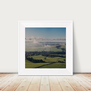 Puede incluir: Una vista panorámica de una campiña verde y ondulada con un pequeño pueblo enclavado en el valle. El paisaje está salpicado de campos, árboles y un camino sinuoso. El cielo es de un azul suave con nubes tenues.