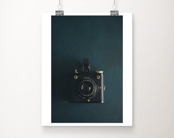 May include: A vintage black and gold camera with the words "Kodak Brownie Flash Six-20" on the front, against a dark teal background.