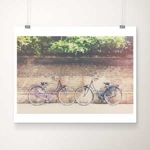Op de afbeelding: Een print met twee vintage fietsen met rieten manden, geparkeerd voor een bakstenen muur en weelderig groen. E&eacute;n fiets is paars, de andere is zwart. De afbeelding heeft een zachte, vintage esthetiek.
