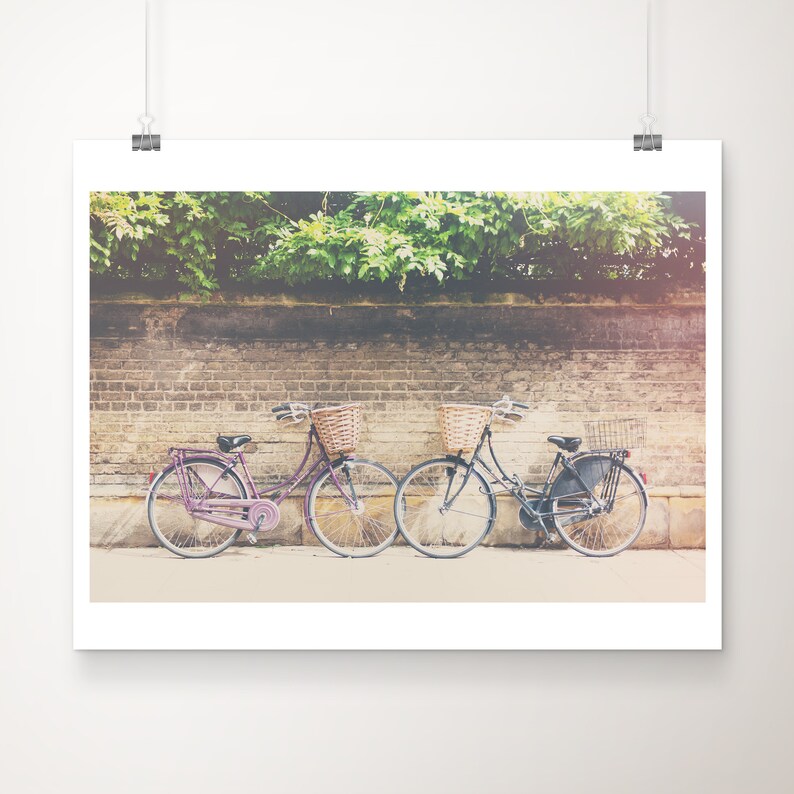 Op de afbeelding: Een print met twee vintage fietsen met rieten manden, geparkeerd voor een bakstenen muur en weelderig groen. E&eacute;n fiets is paars, de andere is zwart. De afbeelding heeft een zachte, vintage esthetiek.