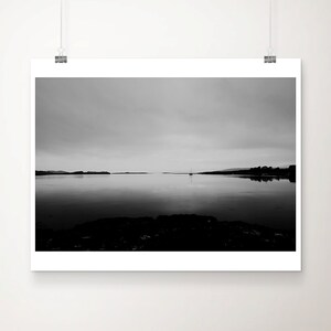 May include: Black and white landscape photograph of a calm body of water reflecting the overcast sky. A small sailboat is visible in the distance, with land on either side. The foreground features dark, textured rocks.
