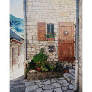 May include: A stone building with a wooden door and a window with shutters. There are potted plants in front of the building and a stone path.